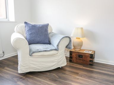 An armchair with a blue cushion and throw next to a wooden box with a lamp on it in a room at 22 Gwel Yr Wyddfa in Menai Bridge
