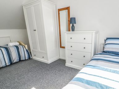 A bedroom with two single beds a white wardrobe a white chest of drawers a mirror and a blue lamp at 22 Gwel Yr Wyddfa in Menai Bridge