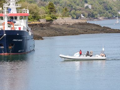 A small boat with three people and a large ship named Prince Madog near a rocky shoreline with trees