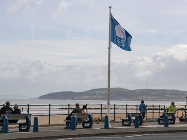 A seaside promenade with benches people sitting and a flagpole on a cloudy day at 22 Gwel Yr Wyddfa in Menai Bridge