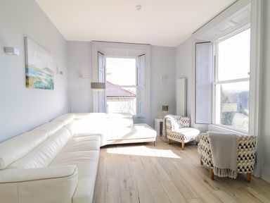 A living room with a white sectional sofa two patterned armchairs two large windows and a floor lamp at Sea Breeze in Ballycastle