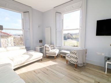 A sitting area with two patterned armchairs and an ottoman near large windows and a white sectional sofa in a room with light wood flooring at Sea Breeze in Ballycastle