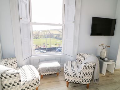 A sitting area with two patterned armchairs and matching ottoman near a large window with a view and a wall-mounted television at Sea Breeze in Ballycastle