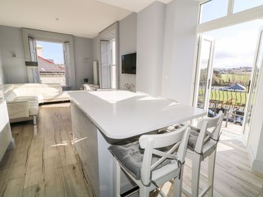 A kitchen island with bar stools and cushions and a living area with a sofa and windows at Sea Breeze in Ballycastle