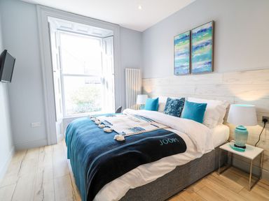 A bedroom with a bed with blue and white bedding beside a window with white shutters and two paintings on the wall at Sea Breeze in Ballycastle