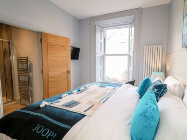 A bedroom with a bed, blue cushions, a window, a wooden door, a wall-mounted TV, and a radiator at Sea Breeze Ballycastle