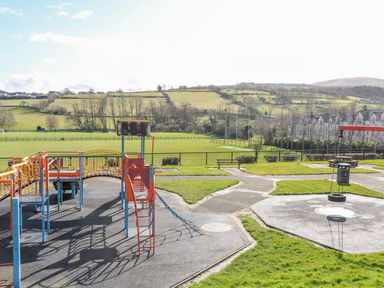 A playground with climbing structures and tire swing overlooking a grassy field and houses in the distance at Sea Breeze in Ballycastle