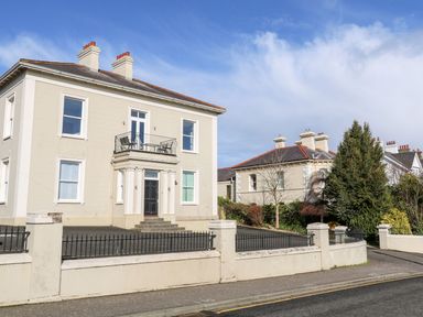A two-story house with a balcony and black front door surrounded by a low wall and trees at Beachcomber in Ballycastle