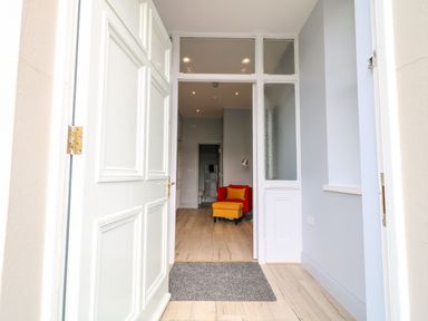 An open white front door leading to a hallway with wooden flooring a red chair with a yellow footstool and a floor lamp Beachcomber Ballycastle