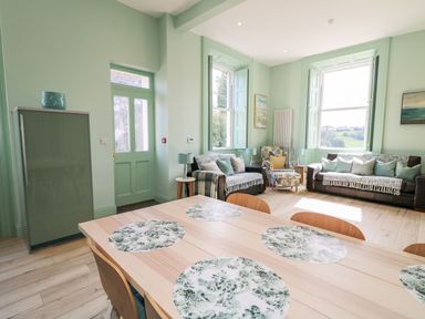 A dining table with six chairs and floral placemats in a living room with two sofas armchair and large windows at Beachcomber in Ballycastle