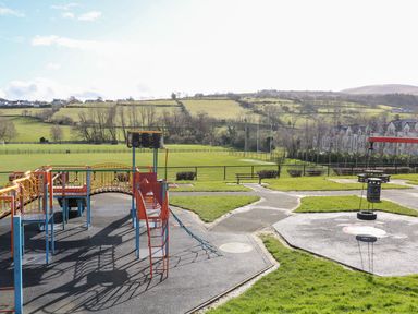 A playground with climbing equipment swing and benches beside a field with hills and buildings in the background at Beachcomber in Ballycastle