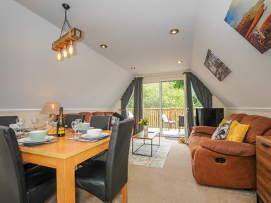 A dining table with place settings and a bottle next to a living area with brown sofas and a sliding door to a balcony at Dart Lodge in Callington