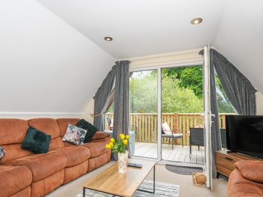 A living room with brown sofas a coffee table with yellow flowers and a TV by a sliding door leading to a balcony with chairs at Dart Lodge in Callington