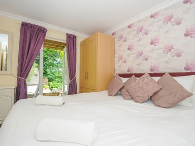 A bedroom with a bed with four pillows and two rolled towels near a window with purple curtains and a wooden wardrobe at Dart Lodge in Callington