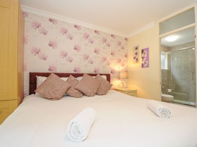 A bedroom with a bed with five pillows two rolled towels a wooden wardrobe a bedside table with lamp and floral paintings with an en suite bathroom at Dart Lodge in Callington