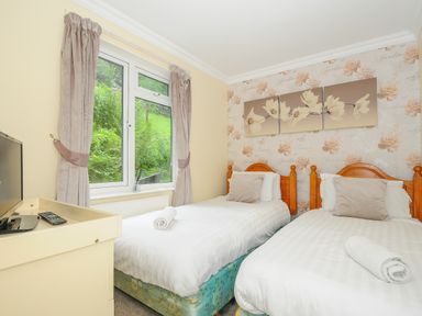 A bedroom with two single beds with white bedding and beige pillows by a window with beige curtains at Dart Lodge in Callington