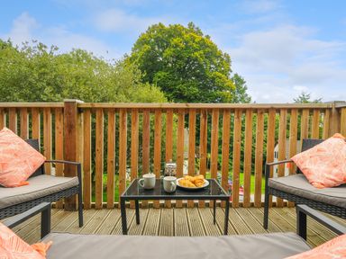 An outdoor wooden deck with a table holding mugs and croissants surrounded by chairs with cushions at Dart Lodge in Callington
