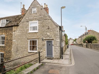 A stone cottage with a gray door and two windows on a corner next to a street with more stone houses at Corner Cottage in Malmesbury