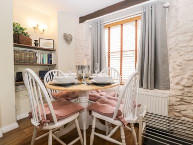 A dining area with a round wooden table and four white chairs with pink cushions at Corner Cottage in Malmesbury