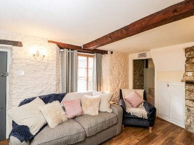 A living room with a gray sofa and an armchair with cushions and a window with curtains at Corner Cottage in Malmesbury
