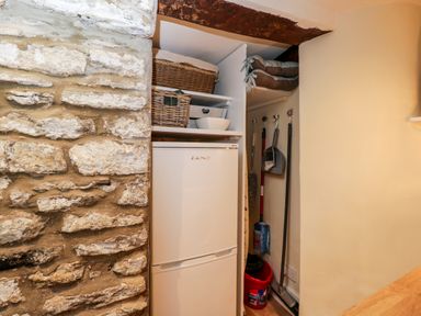 A small storage area with a white fridge freezer baskets cleaning tools and a mop bucket at Corner Cottage in Malmesbury