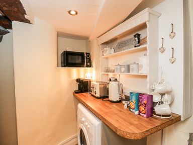 A kitchen corner with a microwave on a shelf a washing machine under a wooden countertop a toaster kettle coffee machine and shelves with mugs canisters and a biscuit box at Corner Cottage in Malmesbury