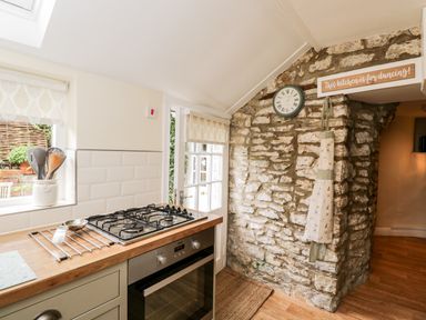 A kitchen with a gas stove oven utensils in a container a clock and an apron hanging on a stone wall at Corner Cottage in Malmesbury