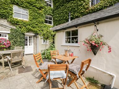 An outdoor patio with wooden tables and chairs surrounded by greenery and flowers at Corner Cottage in Malmesbury