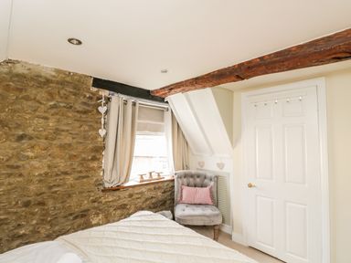 A bedroom corner with a stone wall window with curtains a grey chair with a pink pillow and a white door at Corner Cottage in Malmesbury