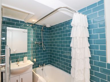 A bathroom with a bathtub shower white ruffled curtain blue tile walls and a white sink at Corner Cottage in Malmesbury