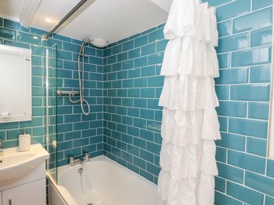 A bathroom with a bathtub shower combination with blue tiled walls and a white ruffled shower curtain at Corner Cottage in Malmesbury