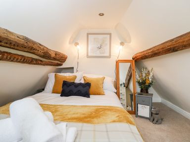 A bedroom with a bed pillows a mirror a bedside table with flowers and exposed wooden beams at Corner Cottage in Malmesbury