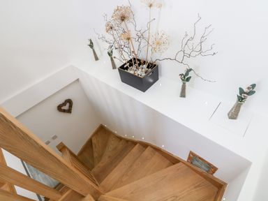 Wooden staircase with white walls decorated with dried plants in vases and a heart-shaped wall hanging at Rock Pool House in Porthleven