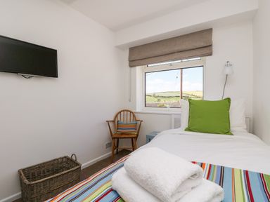 A bedroom with a single bed a green pillow a wooden chair a wall-mounted television and a window showing a countryside view at Rock Pool House in Porthleven