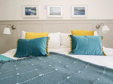 A bed with white sheets blue and yellow pillows and a blue blanket with pictures of ocean waves on the wall at Rock Pool House in Porthleven