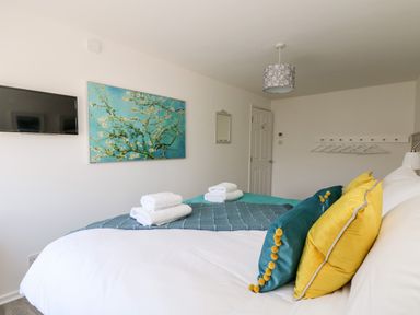 A bedroom with a bed with colorful pillows and folded towels on top a wall-mounted television and a painting at Rock Pool House in Porthleven
