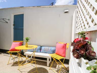 An outdoor patio with a yellow table and two yellow chairs with red cushions and a white bench with blue pillows and plants in a planter box at Rock Pool House in Porthleven