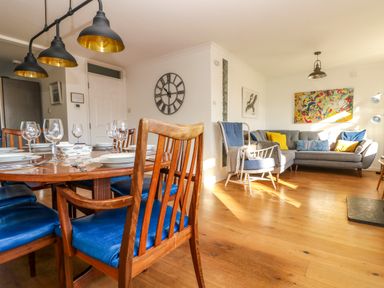 A dining area with a wooden table set with plates and glasses and blue cushioned chairs next to a living room with a grey sofa and an armchair with colorful pillows and wall art at Rock Pool House in Porthleven