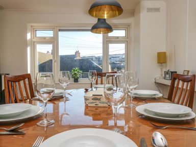 A dining room with a wooden table set with plates wine glasses and cutlery wooden chairs a window with a view and hanging lights at Rock Pool House in Porthleven