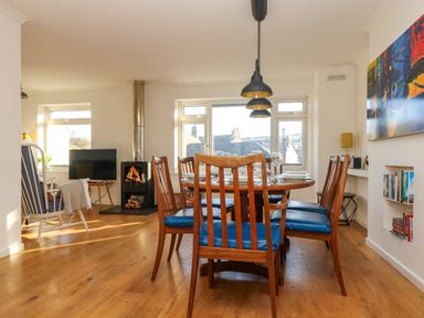 A dining area with wooden chairs with blue cushions around a table and a living area with a fireplace and armchair at Rock Pool House in Porthleven