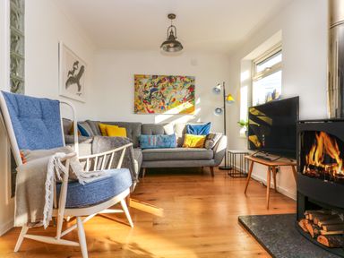 A living room with a blue armchair cushions on a gray sofa a flat screen TV and a wood burning stove at Rock Pool House in Porthleven