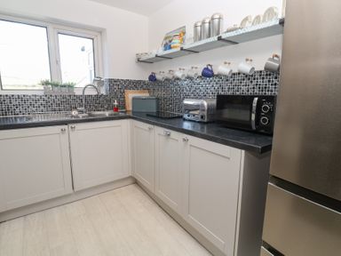 A kitchen with a sink under a window white cabinets black countertop microwave toaster and mugs hanging on a rack at Rock Pool House in Porthleven