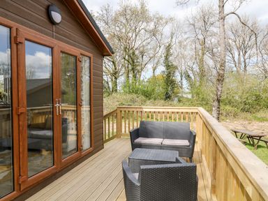 An outdoor wooden deck with black wicker chairs and table outside a building at Dove Lodge in Highampton near Sheepwash