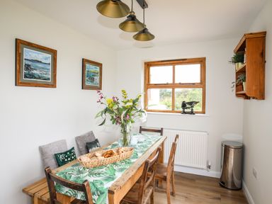 A dining room with a wooden table and chairs a floral table runner flowers on the table two framed paintings on the wall a window with a wooden frame a metal trash bin and a wooden wall shelf with plants at Plas Coch Rhoscolyn in Rhoscolyn