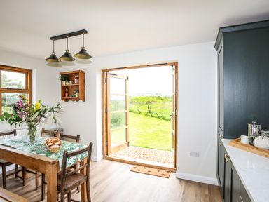 A kitchen dining area with a wooden table and chairs flowers on the table open double doors leading to a grassy garden at Plas Coch Rhoscolyn in Rhoscolyn