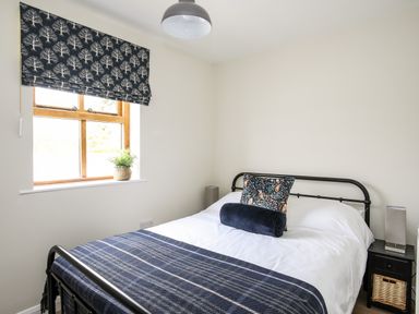 A bedroom with a metal frame bed white bedding patterned cushions and a blue blanket next to a window with a patterned blind and a nightstand with a lamp and basket at Plas Coch Rhoscolyn in Rhoscolyn