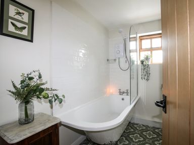 A bathroom with a white bathtub shower and a wooden cabinet with a vase of flowers at Plas Coch Rhoscolyn in Rhoscolyn