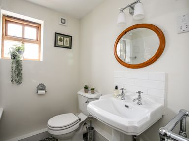 A bathroom with a toilet, sink, round mirror, small window with plants, and framed artwork at Plas Coch Rhoscolyn in Rhoscolyn