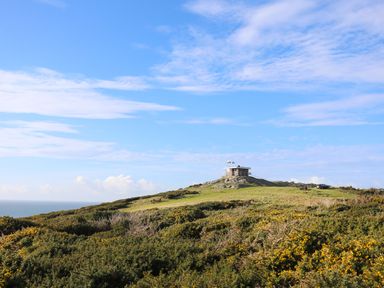 A small building on a grassy hill surrounded by shrubs under a blue sky at Plas Coch Rhoscolyn in Rhoscolyn