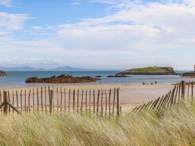 A beach with a wooden fence in front grassy dunes and rocky islets in the sea at Plas Coch Rhoscolyn